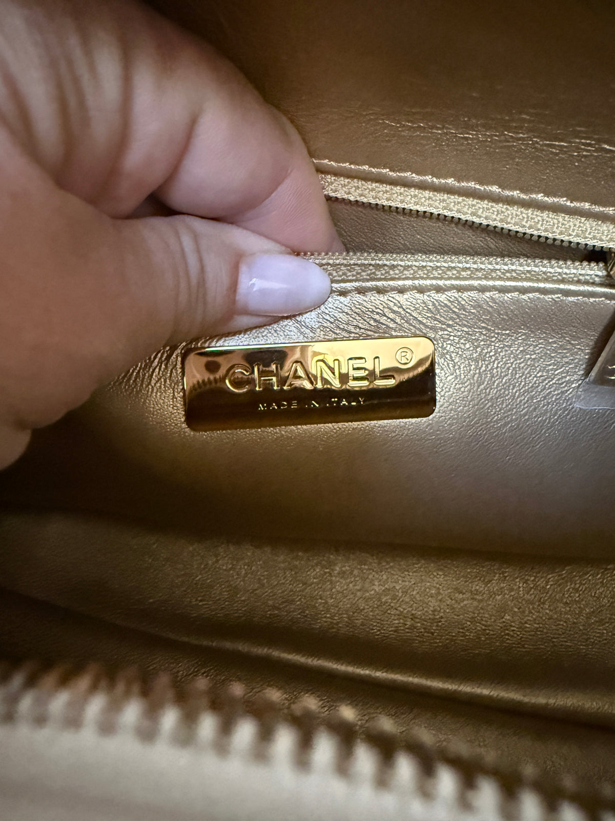 Close-up of a gold-toned Chanel bag interior with a hand pulling a zipper next to a gold metal Chanel logo plaque.