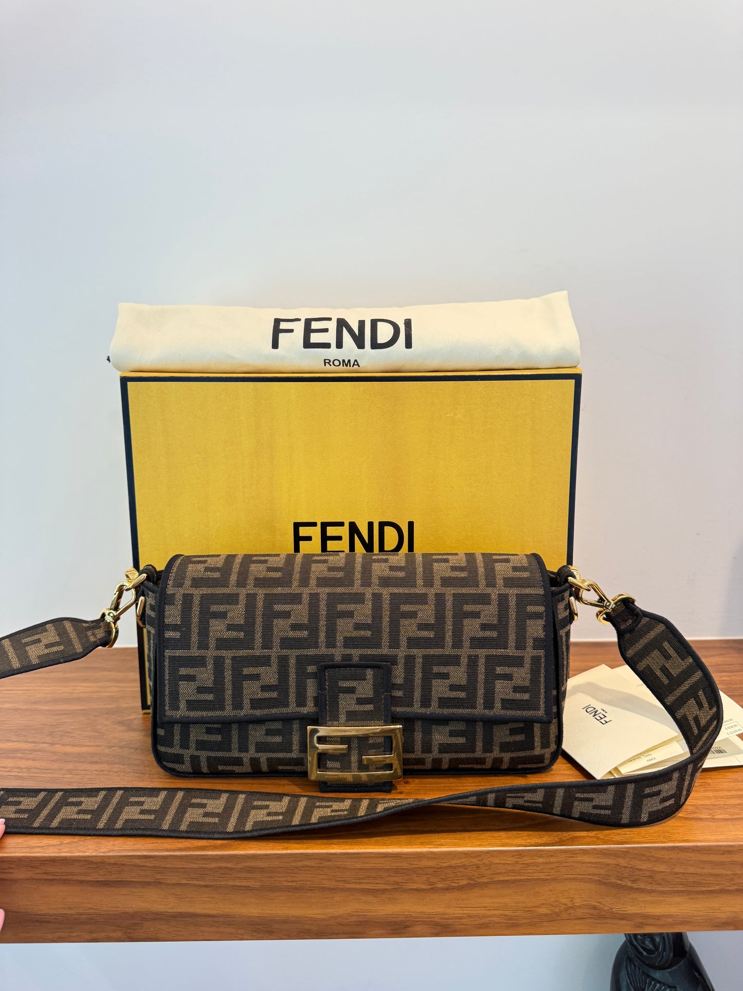 A Fendi Monogram canvas shoulder bag with a gold-tone FF buckle, displayed on a wooden table in front of its yellow and white Fendi packaging box used consignment