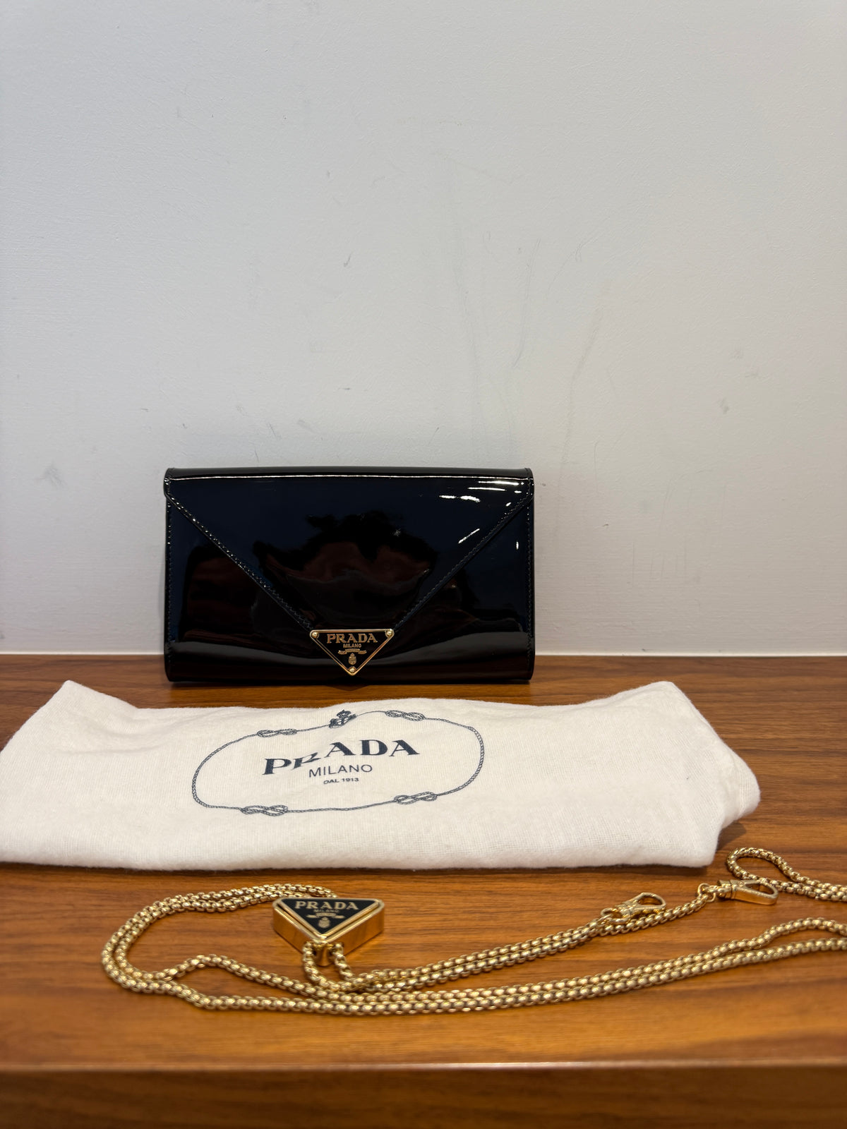 A black patent leather Prada wallet with a gold triangle logo, displayed on a wooden surface with its white branded dust bag and a gold chain accessory.