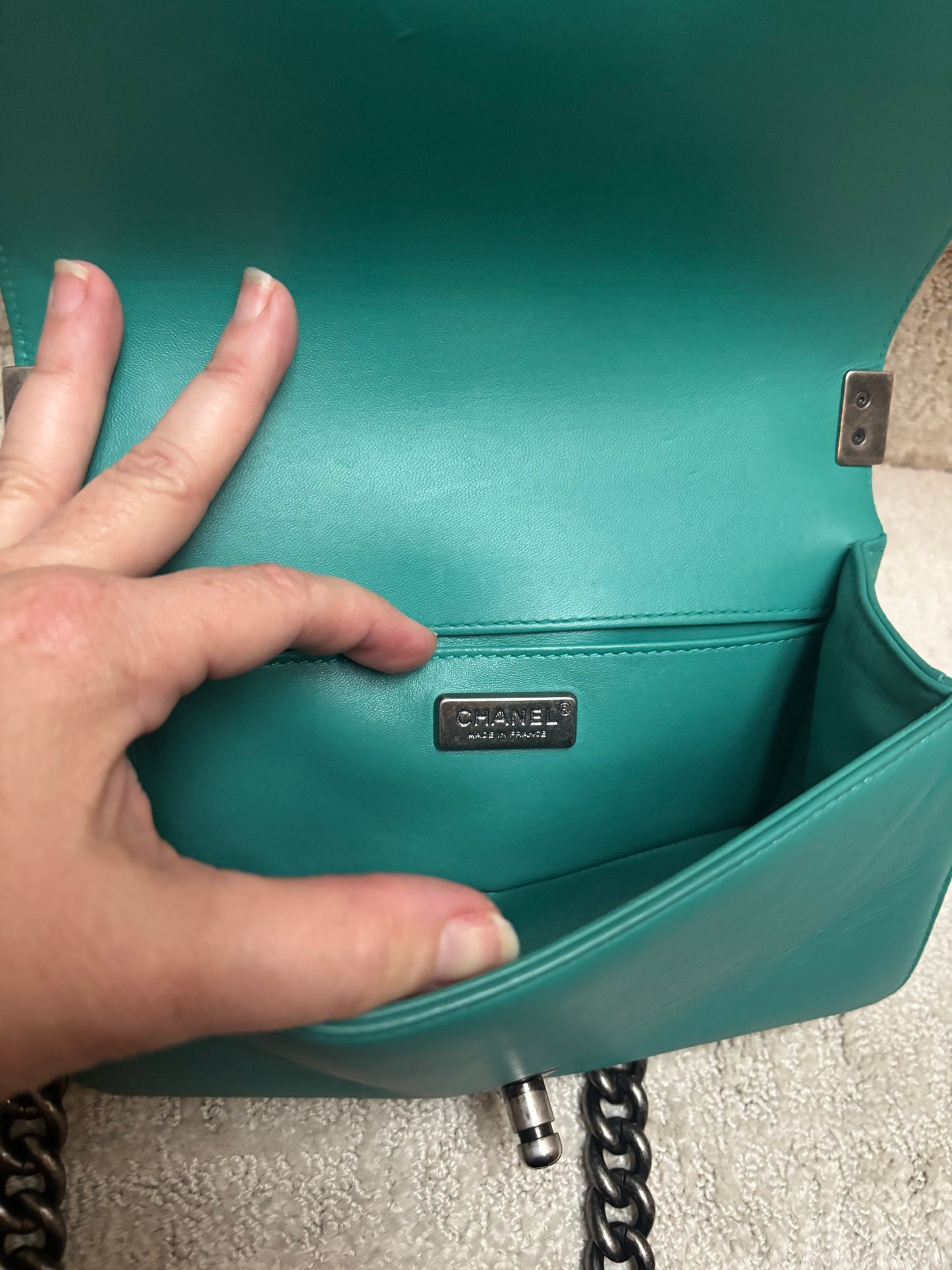 Close-up of a teal Chanel shoulder bag interior with a hand holding the flap open to show the metal logo plaque on the inside pocket for sale from purseonalshopper
