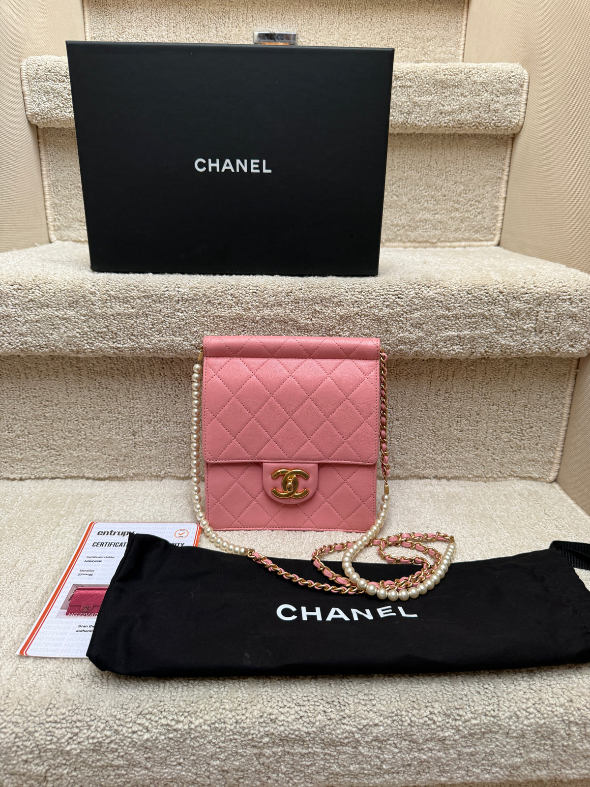 A pink quilted Chanel flap bag with a gold CC turn-lock and chain strap, displayed on carpeted stairs alongside a black Chanel box, dust bag, and paperwork.
