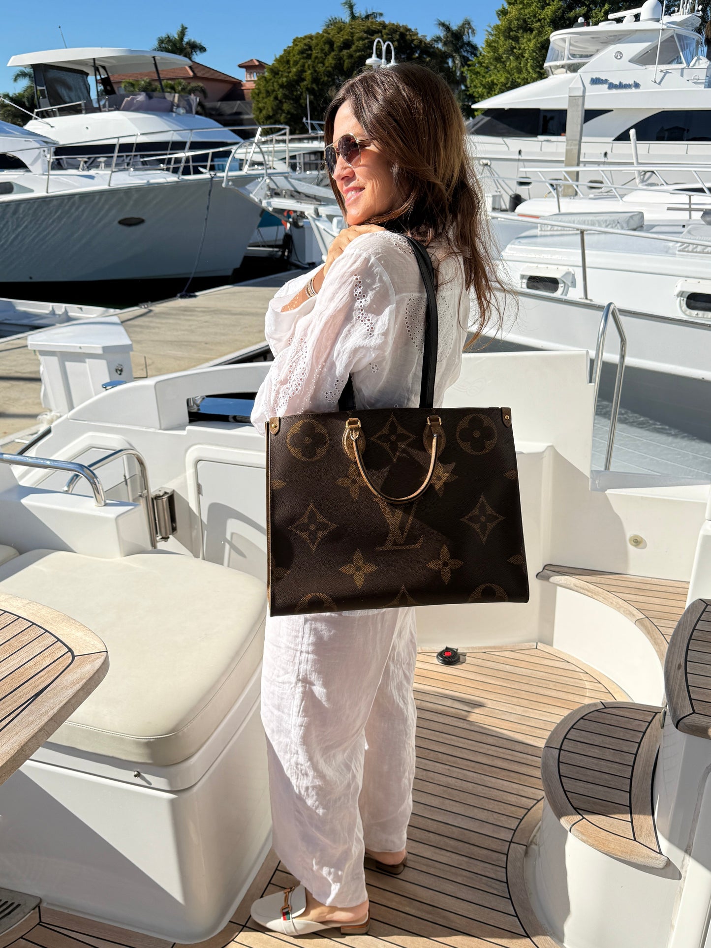 Woman holding a Louis Vuitton on the go GM bag on a yacht deck with a clear blue sky.